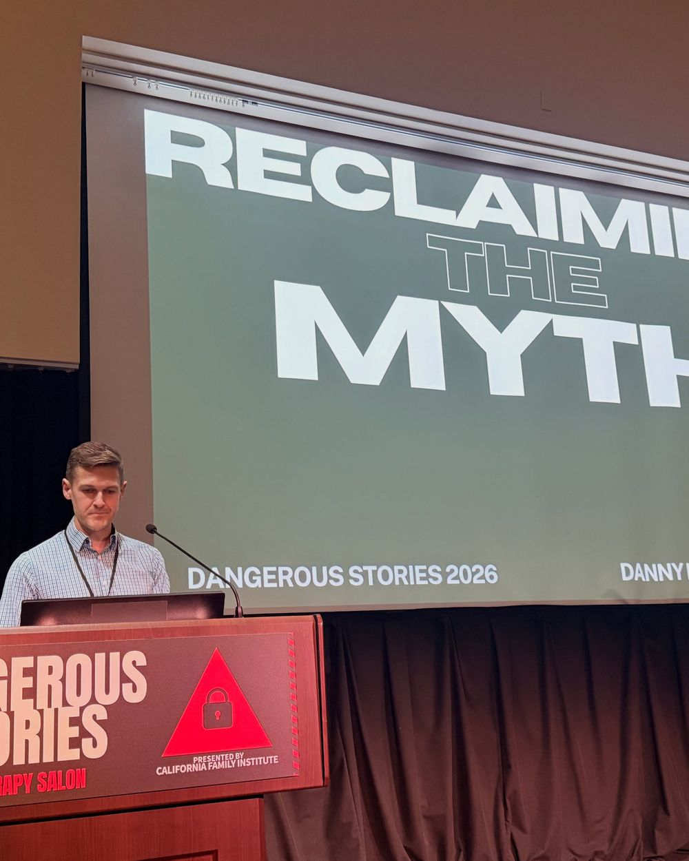Presenter at the lectern under the title slide Reclaiming the Myth